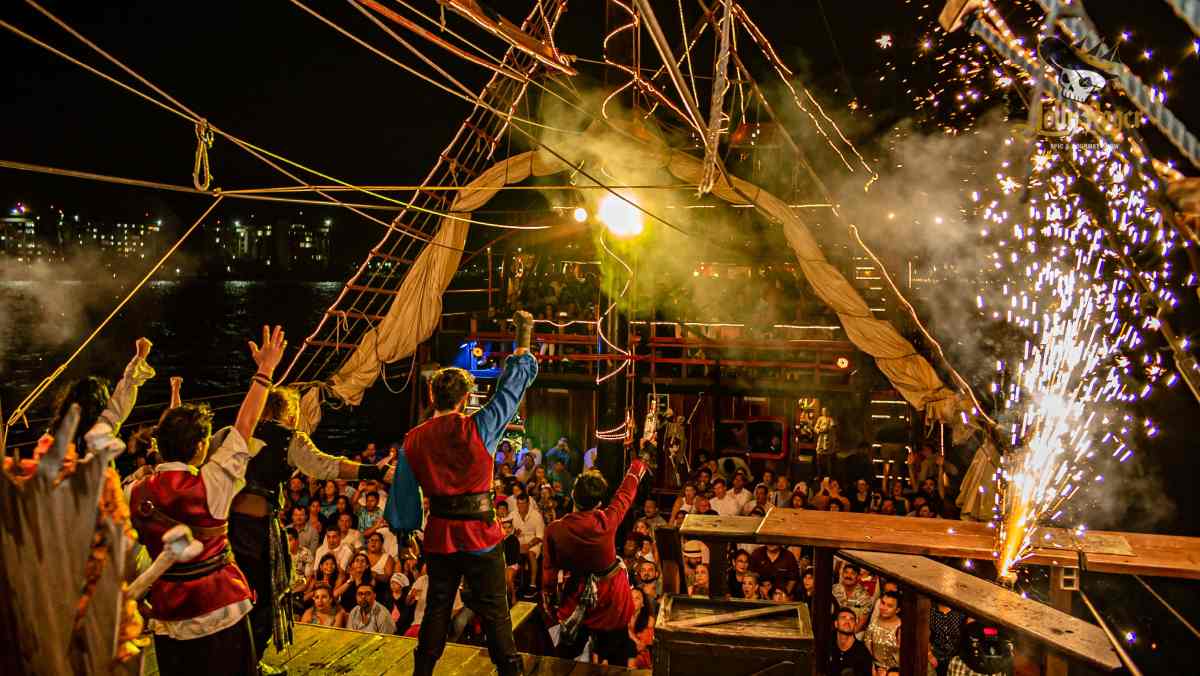 Performers in historical pirate costumes on a Pirate Ship stage entertain an audience at night with fireworks